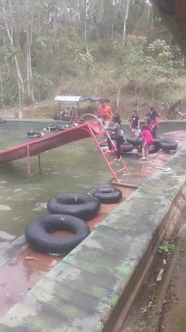 A group of people are enjoying a natural swimming pool. One person is sliding down a red slide into the water. Several inner tubes are visible in and around the pool. Trees and foliage surround the pool area.