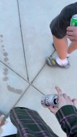 A first-person perspective video shows three people walking on a sidewalk paved with square concrete slabs. Each person is holding a beverage in a can. The person filming is wearing a green plaid skirt and sandals. Another person is wearing black shorts, white socks, and Crocs. The third person is partially visible, wearing black shorts and sandals. There is a trail of liquid spilled on the sidewalk.