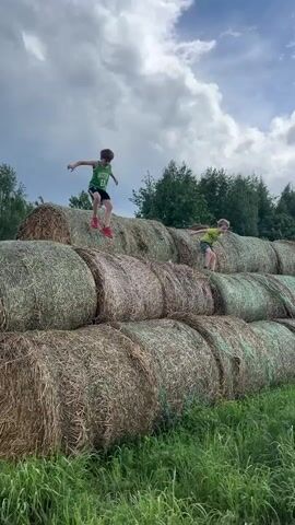 Two boys are walking on hay bales piled in a grassy field. The boy on the left wears a green tank top with black shorts and red shoes. The boy on the right wears a yellow t-shirt and patterned shorts. The hay bales are large, round, and stacked three rows high. Green grass surrounds the hay bales. The sky is cloudy with patches of blue. Trees are visible in the background.