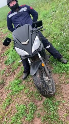 A person wearing a dark blue jacket with red and white stripes, dark pants, and a black helmet sits on a black motorcycle. The motorcycle is positioned on a muddy path surrounded by green grass and some flowers. The person is looking at the camera with their hands behind their back. The video is a close-up shot of the person and the motorcycle.