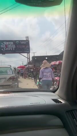 The video captures a street scene from the interior perspective of a vehicle, looking through the windshield. The dashboard, an airbag compartment, and an air vent are visible in the foreground. A large, light-colored sun hat covers the head of a person walking away from the camera in the midground, wearing a light purple shirt with "FOX" printed on the back. The street is bustling with pedestrians and activity, characteristic of a market, featuring various vendors with goods displayed, including green leafy vegetables and round produce in blue tubs. Several red umbrellas provide shade over market stalls. A light-colored sedan is partially visible on the left side of the street. Overhead power lines are strung across the scene. In the background, a large billboard displaying "SUPER NATURAL SHIFT" and images of people stands prominently. Buildings line the street under a bright, partly cloudy sky.