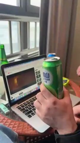 A person holds a green can of soda next to a laptop that is playing a music video on YouTube. The video is for the song "I Got Love" by Miyagi & Эндшпиль featuring Рем Дигга. The person appears to be listening to the music and enjoying a drink in a room with a window overlooking a body of water. There's a green beer bottle and a white mug also on the table.