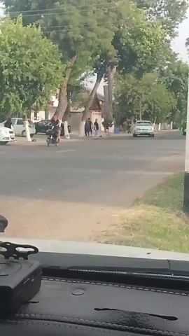 The video captures a street scene from the perspective of a car. In the foreground, the dashboard of the car is visible. The road ahead has several vehicles, including a white car turning, and other cars driving by. On the sidewalk and crossing the street, there are several people, some appearing to be walking and others interacting. Trees line the street, and buildings are visible in the background. The scene appears to be an ordinary street in a residential or commercial area.