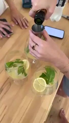 The video shows a person making cocktails at a wooden table. Three large stemmed glasses filled with a yellow liquid, ice, mint sprigs, and lemon slices are on the table. The person is pouring a liquid from a small green bottle into a stainless steel jigger. Another person’s hands are partially visible on the left side of the frame. A smartphone, a black speaker, and a plastic soda bottle are also on the table.
