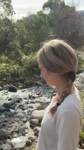 A woman with blonde hair in a braid, wearing a white long-sleeved shirt, stands outdoors near a rocky stream. She holds a black object, which appears to be a gun, in her right hand and aims it towards the water. The environment is natural, with trees and vegetation surrounding the stream. The lighting suggests it is daytime.