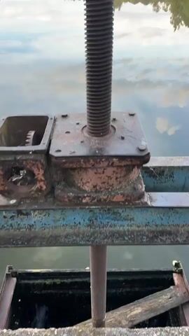 The video opens with a close-up of a large, rusted metal screw mechanism with visible threads, likely part of a gate or dam control system. The screw is positioned on a metal base that appears weathered. Beyond this mechanism, a calm body of water stretches out, reflecting the sky above. The water is a soft blue, mirroring scattered white clouds.

As the video progresses, the camera pans across a serene lake or pond. The reflections on the water are striking, with the sky and the treeline on the opposite bank creating a near-perfect mirror image. The shoreline is densely populated with various trees, including evergreens and deciduous trees, forming a lush green border. In the distance, the landscape appears gentle and undulating, with more trees and some hints of open sky.

The sky is a mix of blue and white, with wispy clouds and some more substantial formations. The overall impression is one of a tranquil, natural setting, possibly in the early morning or late afternoon due to the soft light and the presence of what might be a light mist or fog over the water. The scene evokes a sense of peace and quiet. There is no visible text in the media file. The location cues (Пекалінскі сельскі Савет, Belarus) suggest a rural or natural area in Belarus.