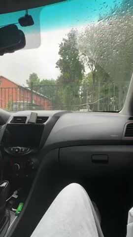 The video is shot from inside a car, looking out the windshield on a rainy day. The dashboard of the car is visible, including the steering wheel, center console with a screen, and air vents. A phone is mounted on the dashboard. Outside, trees and a fence are visible, along with a brick building. Water droplets are visible on the windshield, distorting the view slightly. A person's leg is visible in the lower right corner of the frame, suggesting the videographer is seated in the car.
