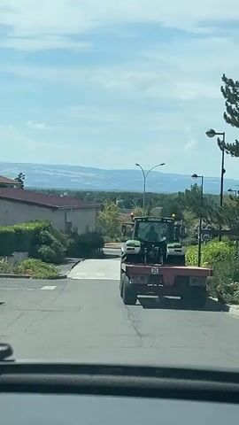 A tractor with a trailer attached is driving away from the camera on a paved road. The tractor is green and white and has a person inside. The trailer is red and appears to be empty. There are buildings and trees on either side of the road, and a landscape with hills in the background. The sky is blue with some clouds. The video appears to have been recorded from a moving vehicle.