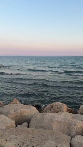 A slow pan reveals a calm ocean with gentle waves lapping against a rocky shore. The sky transitions from a soft blue at the top to delicate shades of pink and orange near the horizon, indicating either sunrise or sunset. Large, light-colored rocks form the foreground, with smaller stones and pebbles visible at the bottom. In the distance, a line of submerged rocks creates a subtle breakwater. The overall atmosphere is peaceful and serene.