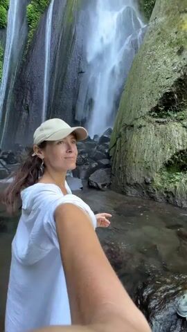 A woman in a white hoodie and a beige baseball cap takes a selfie video in front of a waterfall. She is positioned on the left side of the frame, with the waterfall and a moss-covered rock formation behind her. The water cascades down a rocky cliff, and lush green vegetation surrounds the area. The woman smiles at the camera as she extends her arm, holding her phone out to capture the shot. Two pairs of sandals are visible on a rock to the right of the frame.