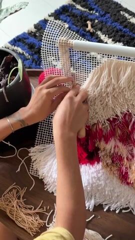 The video shows a close-up overhead view of a person's hands working on a textile art piece. The person is using a tool to push strands of yarn through a white mesh canvas that is stretched on a frame. The artwork itself is a textured piece with various colors of yarn, including white, cream, brown, and red, with some fringe elements visible. Several bundles of yarn are lying on the wooden surface below the canvas. In the background, a dark rug with blue and black stripes is partially visible, along with a dark pot containing a plant. The lighting suggests an indoor setting with natural light coming from the right. The person's hands are the primary focus, demonstrating the crafting process.