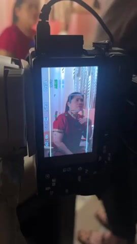 A medium shot shows a woman in a red outfit sitting on a chair, being filmed. The camera is positioned in front of her, and we see the recording screen with a reflection of the woman. In the background, there is a macrame wall hanging and a wooden ladder with a clock. A person's arm and shoulder are visible in the foreground, holding a phone. The woman gestures with her hand as she speaks. The scene appears to be an indoor setting, possibly a room or a studio.