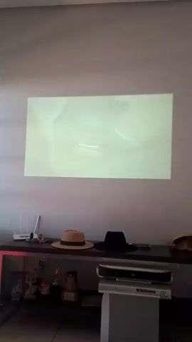 A static, eye-level shot of a projector screen on a light-colored wall is shown. The screen displays a logo featuring a striped circle above the word "MORENA." Below the projected image, a dark shelf holds various items including a white router with antennas, several trophies or decorative objects, a straw hat, a black fedora, and a silver stereo system. At 0:03, the projected image abruptly disappears, leaving the wall blank, while the camera remains still. The lighting in the room is dim.