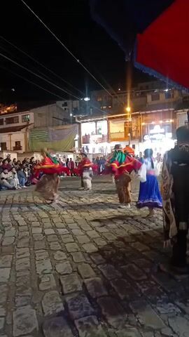 The video captures a lively street performance at night. Several dancers, some in red and fur-like costumes with green accents, and one woman in a blue skirt, are performing on a cobblestone square. A crowd of onlookers surrounds the performance area, some seated and others standing. The square is illuminated by streetlights, casting a warm glow on the scene. Buildings form the backdrop, with some featuring balconies and visible interior lighting. A large red umbrella with blue trim partially obscures the upper right corner of the frame, and a person wearing a patterned shawl stands in the foreground on the right side of the video.