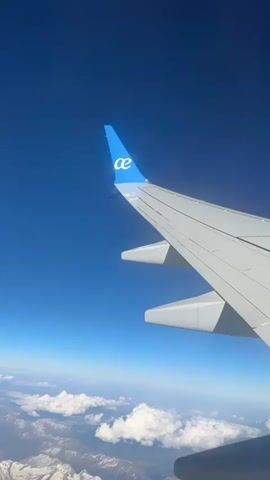 The video shows a view from an airplane window, specifically the wing of the plane with the "ae" logo on the wingtip. The sky is a clear blue. Below the plane, there are clouds and a landscape of snow-capped mountains. The camera is relatively stable, providing a clear view of the scenery.