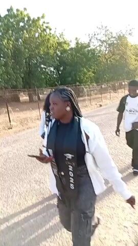 The video depicts three young individuals walking along a gravel road. One is a young woman with braids, wearing a white jacket over a black top and gray pants, holding a phone. Another is a young man in a white shirt with black accents, black pants, and sneakers. The third person is a young woman wearing a white blouse, black pants, and a graduation scarf that says "Class of 2025." She carries a black purse. The young man is seen smiling and appears to be playfully posing. The scene is outdoors with trees and a fence visible in the background under a bright sky.