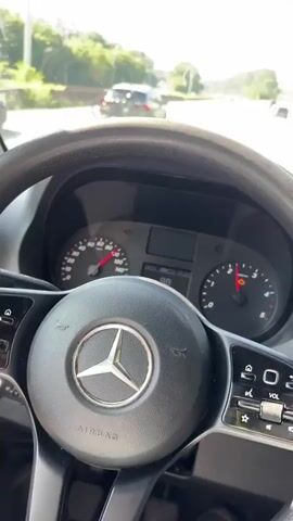 The video shows a first-person view from inside a vehicle, likely a van, driving on a highway. The driver's side dashboard and steering wheel are visible in the foreground. The steering wheel has a Mercedes-Benz logo. The instrument cluster shows the speedometer needle around 80-90, and a warning light is illuminated. The road ahead is clear with other vehicles in the distance. Trees and greenery line the sides of the highway, and the sky is blue with some light cloud streaks. The vehicle appears to be moving at a moderate speed on a sunny day.