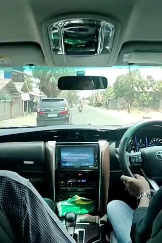 The video is filmed from the backseat of a car, looking forward through the windshield. The dashboard of the car is visible in the foreground, with a central touchscreen display showing information. A person's hands are visible on the steering wheel, indicating they are driving. Another person's legs are visible in the lower left corner of the frame. Outside the car, a street scene is visible, with other vehicles, buildings, and trees lining the road. The weather appears to be sunny. The car is moving forward on a paved road.