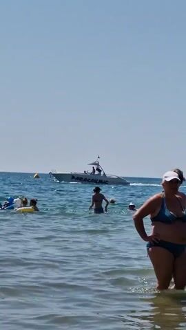 The video shows a sunny day at a beach with people in the water and a boat in the distance. In the foreground, a person in a blue bikini stands in the shallow water, facing away from the camera, with their hands on their hips. Further out in the water, several other people are visible. Some are swimming, while others are playing with inflatable toys, including a yellow ring and a blue and white inflatable animal. In the background, a white motorboat with "PARASAILING" written on its side is moving across the blue ocean. There are a few people on the boat, including one standing near the stern. The sky is clear and blue.