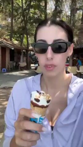 A woman in sunglasses and a purple jacket is eating an ice cream cone. She is standing outside on a sunny day. Behind her are trees, beach houses, and some tables and chairs. She takes a bite of the ice cream cone, chews, and looks at the camera.