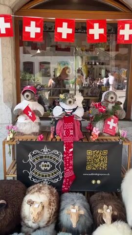 The video shows a shop window decorated with Swiss flags. In the foreground, three teddy bears are displayed on a wooden stand. The teddy bear on the left is wearing a white shirt with a Swiss flag and a red hat. The middle teddy bear is dressed in a traditional Swiss outfit with a flower crown and is holding a red stocking with white patterns. The teddy bear on the right is wearing a grey outfit and holding a red bag with a white Swiss cross. The stand also has a black banner with a logo and a QR code. Below the stand, several sheep decorations are visible. In the background, through the shop window, several people can be seen browsing inside the store. The lighting suggests it is daytime.