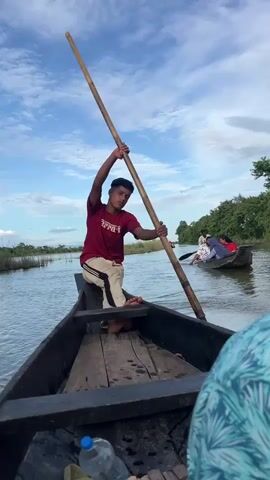 The video shows a young man in a maroon t-shirt and white pants steering a wooden boat with a long pole. He is sitting in the front of the boat, facing forward. The boat is on a river, and there are other boats in the background. One boat has a few people in it, including a woman in a blue headscarf. Another boat appears to be a motorboat. The river is surrounded by lush green trees and vegetation. The sky is blue with some white clouds. The water is calm, and the scene appears to be peaceful. The camera is positioned low, giving the viewer a sense of being in the boat with the steersman.