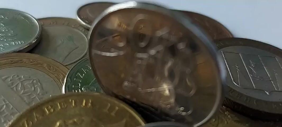 The video shows a close-up, high-angle view of a pile of assorted coins. The coins are varied in size, color, and apparent origin, with some appearing to be gold-colored, others silver-colored, and some with a coppery hue. The surface they are resting on is a plain, light-colored background, likely a table or similar flat surface. The coins are arranged in a loose, overlapping manner, creating a textured visual. Some coins are in sharp focus, revealing details of their inscriptions and imagery, while others are blurred due to the shallow depth of field. In the foreground, a gold-colored coin with the profile of Queen Elizabeth II is visible. To its right, a silver-colored coin with a design that appears to be a ship is prominently displayed. Other coins with visible text include one with "ELIZABETH II" and another with "1987". The lighting is bright and even, creating subtle reflections on the metallic surfaces of the coins. There are no people or discernible actions taking place, the focus is solely on the static arrangement of the coins.