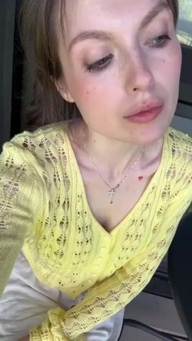 A fair-skinned woman with light brown hair pulled back in a loose ponytail is the subject of a close-up video. She has a small red heart tattoo on her chest and wears a silver necklace with a cross pendant. Her top is a pastel yellow, knitted cardigan with an intricate openwork pattern. The woman opens her eyes, glances to the right, smiles faintly, and then looks upwards. The background is blurred, but hints of a window and a curtain are visible.