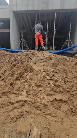 A person in an orange construction outfit is standing on top of a mound of dirt. They are using a tool to knock against a metal pole. Behind them, a concrete structure is visible with several more metal poles supporting a wooden formwork. Blue pipes run along the ground in front of the dirt mound.