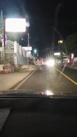 The video shows a view from inside a car driving on a street at night. The street is paved and has yellow lines marking the lanes. There are buildings on both sides of the street, some with signs and lights. Several other vehicles are on the road, with their headlights on. There are streetlights illuminating the area. The car is moving forward, and the perspective is from the driver's seat.