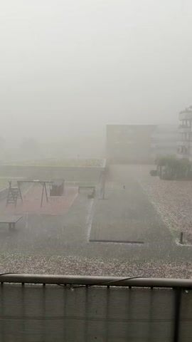 The video shows an outdoor courtyard with buildings and a playground under heavy snowfall. The visibility is poor due to fog or heavy snow. The ground is covered with a thick layer of snow, and snowflakes are visible falling. There are no people visible in the video. The objects in the scene include apartment buildings, a playground with swings and a slide, a table tennis table, benches, and a rooftop garden with some greenery. The environment is a courtyard between buildings, likely in a residential area. The weather conditions are severe winter weather.