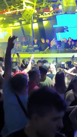 Here's a concise description of the video:

The video captures a vibrant scene inside a nightclub. A large crowd of people is dancing with their hands raised in the air. Above the crowd, on a raised platform, a group of DJs are performing. A large screen behind the DJs displays colorful graphics and the word "TERMINAL" is visible. The club is lit with bright, colorful lights, including yellow and blue beams that sweep across the crowd. Wires and fixtures are visible on the ceiling.