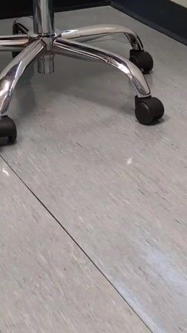 The video shows the base of a chrome office chair with black wheels on a gray tiled floor.  A small, chrome object sits on the floor to the right of the chair. The bottom of a dark wall runs along the top of the frame.