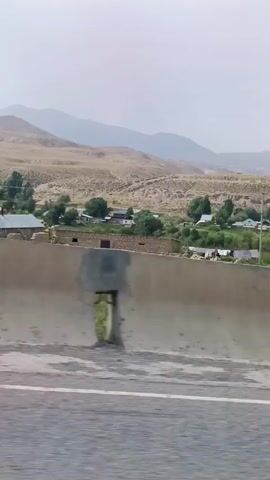 This is a video taken from a moving vehicle, likely a car, showing a rural landscape. The camera is positioned to capture the view from the side window. In the foreground, there is a concrete barrier, possibly a highway divider, with a gap or opening visible in it. Beyond the barrier, there is a grassy area, followed by a cluster of buildings that appear to be houses or small structures with pitched roofs. Some of these buildings have windows and doors. Further in the background, there are more buildings scattered amongst trees and vegetation. The terrain behind the settlement is characterized by rolling hills or mountains with dry, barren slopes, possibly indicative of a desert or semi-arid region. The sky is overcast and appears hazy. There are also some utility poles with wires visible in the scene. The overall impression is of a quiet, rural area with a modest settlement nestled within a vast, undeveloped landscape.