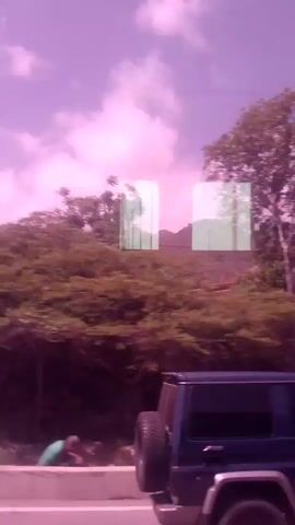 This is a short video clip, filmed from the perspective of a moving vehicle, likely traveling along a coastal road. The scene captures a mix of natural and urban elements. Lush green trees, some appearing to be palm trees, line the road, interspersed with foliage and some bare patches of earth.

Several buildings are visible, ranging from a small, older house with a stone pathway leading to its entrance, to a tall, modern apartment or office building with many windows. Another building, appearing to be a hotel, is visible in the distance. The presence of a black SUV with a spare tire mounted on the back suggests ongoing transportation or travel. The road itself appears to be paved, with white lane markings visible, indicating it's a public thoroughfare.

The overall impression of the scene is bright and sunny, suggesting a daytime setting. The sky is filled with clouds, hinting at potentially dynamic weather, but the light suggests it's not heavily overcast. The filtering of the video gives the scene a pinkish hue, which might be an effect of the camera or lighting conditions, making the sky and some surfaces appear with this color cast.

No people are directly visible in the clip, but the presence of vehicles and buildings implies human activity and habitation in the area. The setting appears to be a developed area, likely a town or city on the coast, given the vegetation and proximity of buildings to the road. There are no discernible signs with readable text within the frame. The activity captured is simply the movement through this environment, offering a glimpse of the surroundings.