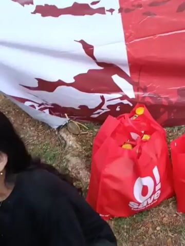 The video shows a close-up of a large red and white banner, likely a flag or banner for an event. The banner has text on it that reads "DIRGAHAYU REPUBLIK INDONESIA" and "17 AGUSTUS 1945 - 2026". Below this text, it says "Warga RT 01 RW 08 Cluster Arcadia - Forest Hill". There are also red shopping bags filled with yellow objects, possibly food or snacks, placed on the ground in front of the banner. The ground appears to be covered in grass and some scattered leaves or debris. The lighting suggests it is daytime. The video is shot from a low angle, looking up at the banner.