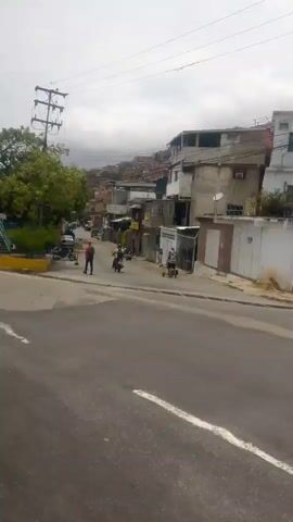 The camera moves along a road, showing the side of a green pedestrian bridge over the road, a colorful fence around a basketball court, trees, and buildings on a hillside in the background. A red bus with "GOB La Guaira" and "029" on the back drives past. People stand near the bus. The sky is overcast.