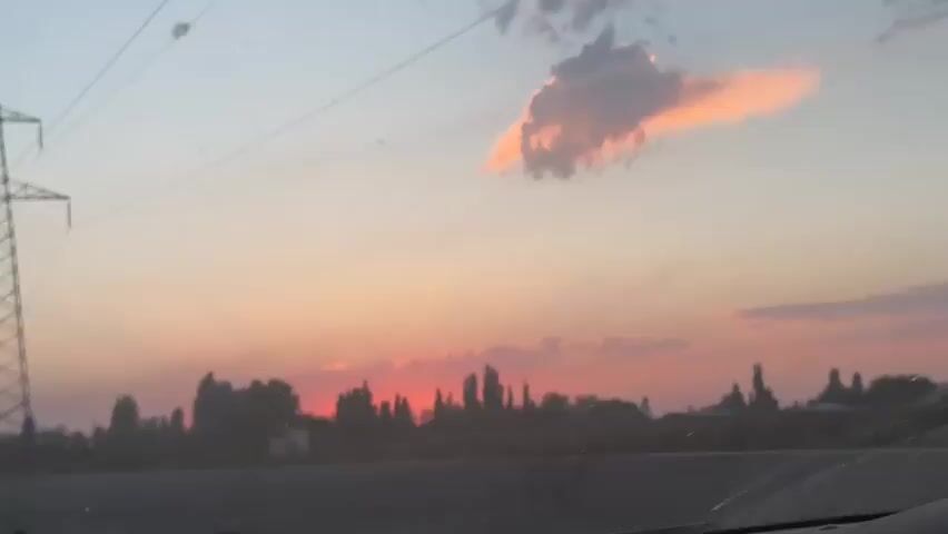 A power tower stands tall on the left side of the frame, silhouetted against a sky painted with the soft hues of sunset. Wispy clouds catch the fading light, tinged with orange and pink. Below the sky, a line of trees forms a dark silhouette, suggesting a landscape of rural or suburban development. The sun itself is a small, fiery orb just beginning to dip below the horizon, casting a warm glow through the trees. Power lines stretch across the frame, connecting to the tower and disappearing out of view. The overall impression is one of a tranquil evening scene, marked by the presence of man-made infrastructure against the backdrop of natural beauty.