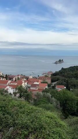 The video shows a coastal town with red-tiled roofs and lush green vegetation overlooking a calm blue sea. Several boats, including a sailboat with a tall mast, are anchored in the bay. A small island is visible in the distance. The sky is overcast with a mix of clouds. The camera slowly zooms in on the boats.