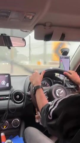 This is a first-person perspective video taken inside a car as it is being driven in the rain. The camera is positioned to show the driver's hands on the steering wheel, the car's dashboard, and the windshield. The driver is wearing a watch and a jacket with white stripes on the sleeves. A navigation app is displayed on a phone mounted on the dashboard. The windshield wipers are active, clearing rain from the windshield. Outside the car, through the windshield, a road can be seen with yellow directional signs indicating a curve to the left. The weather appears overcast and rainy.