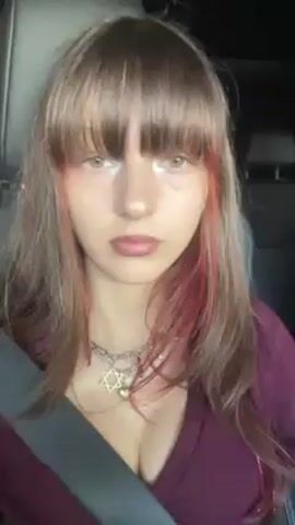 A young woman with light brown hair, bangs, and red streaks is in the passenger seat of a car. She's wearing a maroon top and has a silver necklace with a Star of David pendant. She looks at the camera and smiles. The camera angle is slightly tilted. She is wearing a seatbelt.
