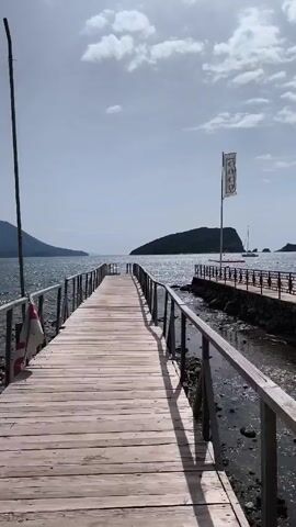 The video shows a straight wooden pier leading out into a body of water, likely the sea. The pier is lined with metal railings on both sides. The water is choppy and the sky is overcast with some patches of blue visible. In the distance, there is an island and a few boats. To the right of the pier, there is another, shorter pier made of brick with similar railings. A flag with the word "COCO" printed vertically is visible on the shorter pier. There are no people visible in the video.