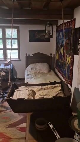 The video pans across a rustic room, possibly a bedroom or living space, with a wooden floor and exposed wooden ceiling beams. A small, antique-looking cradle hangs from a rope suspended from the ceiling, draped with a patterned blanket and pillows. To the right of the cradle is a single bed with a wooden headboard, covered with white bedding and a large pillow. Above the bed, a kerosene lamp hangs from the ceiling. On the wall to the right of the bed, a colorful, woven tapestry is displayed. A window with wooden panes is visible on the back wall, showing greenery outside. To the left of the window, a small table holds a few items. Further to the left, a rocking horse with a patterned coat sits near a wooden chair. In the foreground, a long wooden table with a blue-painted base is visible, set with two cups and a jug. A sewing machine and other small items are also on this table. A rug with a geometric pattern is laid out on the floor. The room appears to be illuminated by a overhead light fixture and natural light from the window.