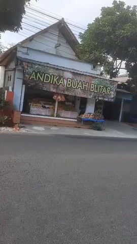 A video captures a street view in front of a fruit stand named ANDIKA BUAH BLITAR. The fruit stand is a small building with a banner displaying its name. Various fruits are displayed in front of the stand, including what appears to be durian on the right side. Behind the fruit stand, there are houses and trees. On the street in front of the stand, a flatbed truck is parked, loaded with sacks, and a motorcycle is visible in the background. The sky is clear and bright, suggesting it is daytime. The video appears to be a continuous shot with a slight pan to the right.