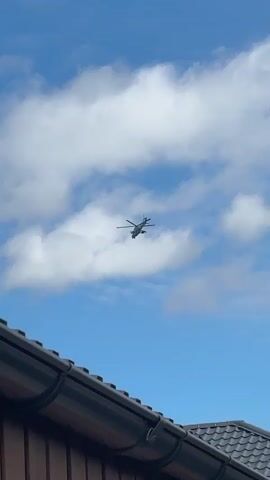 A helicopter flies in the sky. The helicopter is dark gray and has a rotor on top. Below the helicopter is a blue sky with white clouds. In the foreground, part of a dark brown house roof is visible.