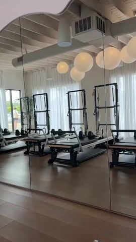 The video displays the interior of a bright and modern pilates studio, characterized by a spacious layout and clean design. The dominant feature is a large mirrored wall reflecting the room, creating an illusion of extended space.

In the reflected view, several pilates reformers with attached towers are neatly arranged on a wooden floor. The reformers are black with natural wood accents on their sliding carriages. Above, the ceiling features exposed white wooden beams, recessed lighting, and rectangular air conditioning vents. Numerous white, spherical paper lanterns of varying sizes hang from the ceiling, adding a decorative and soft lighting element.

Behind the reformers, large windows are covered with sheer white curtains, allowing ample natural light to fill the room, suggesting it is daytime.

Initially, the studio appears empty, but then a person, a woman with blonde hair, appears in the reflection. She is seated cross-legged on one of the pilates reformers, wearing dark athletic attire. She is holding a pink-cased smartphone up, seemingly taking a selfie or recording herself. A green water bottle rests on the floor next to her reformer.

The overall atmosphere is calm and well-lit, indicating a professional fitness environment in Solana Beach, United States of America. No specific text is clearly legible within the video frame.