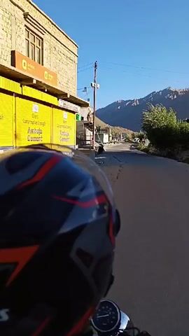 The video is a first-person perspective shot from a motorcycle helmet as the rider travels down a paved road in a mountainous, rural area. The rider is wearing a black, red, and gray helmet with the brand "GLIDERS" visible. Buildings with stone and painted facades line the left side of the road, with one building featuring a bright yellow storefront. In the background, majestic mountains rise under a clear blue sky. The road appears to be relatively empty, with a few parked vehicles and distant structures visible. The overall impression is one of a scenic journey through a remote landscape.