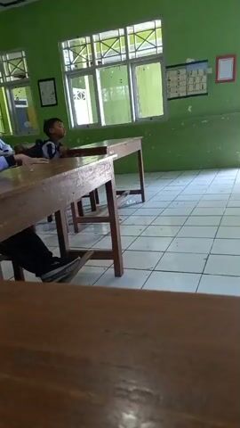 A boy stands in a doorway, looking back into a classroom. The classroom has green walls, tiled floors, and wooden desks. Windows are visible on one wall. Another boy is sitting at a desk, looking forward. He is wearing a backpack and a jacket with a striped pattern. Other students are visible in the background. The boy in the doorway is wearing dark pants and a jacket with a striped pattern. He appears to be talking or yelling.