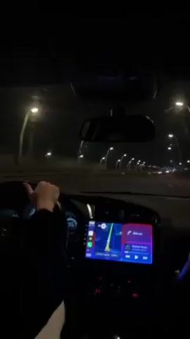 The video shows a car driving on a highway at night. The driver's hand is visible on the steering wheel. The car's dashboard is lit up, displaying a GPS navigation screen and music information. Streetlights line the highway, and other cars can be seen in the distance.