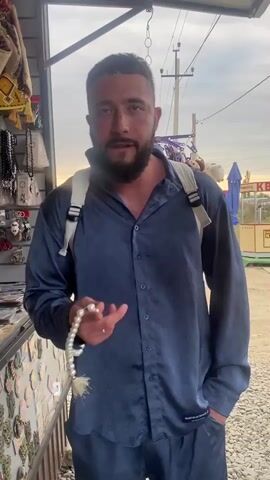 A man with a beard stands in front of a stall, holding prayer beads. He wears a blue patterned shirt and backpack. The stall appears to be selling souvenirs. The background shows a street with a utility pole and trees. The man is looking directly at the camera.